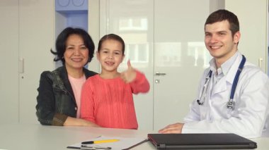 Neşeli küçük bir kız, parlak bir klinikte, destekleyici annesinin ve dost canlısı bir erkek doktorun yanında baş parmağını sallıyor. Bu iç açıcı sahne başarılı bir pediatrik ziyaretin zevkini yakalıyor, sağlık hizmetlerindeki güven ve bakım bağını gözler önüne seriyor.