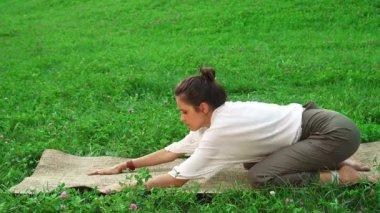 Siyah saçlı, at kuyruklu genç kadın, yemyeşil çimlerin arasında doğal bir minder üzerinde zarif yoga pozları veriyor. Huzurlu ifadesi, dinleyicileri sağlıklı bir yaşam tarzına ve sakinleştirici özüne davet ediyor.