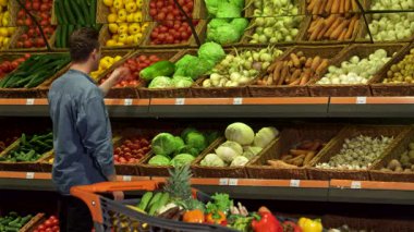 Şık, sarışın bir adam hareketli bir süpermarketin üretim bölümünde canlı bir Napa lahanasını inceliyor. Taze sebze ve meyve çeşitleriyle çevrelenmiş sağlıklı yaşam ve dikkatli alışverişin özünü somutlaştırıyor.