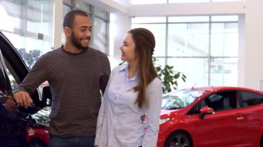 Satıcıda yeni araba alımlarını kutlayan mutlu ırklar arası bir sevinç ışıltısı. Yakışıklı Afrikalı Amerikalı adam gururla arabanın anahtarlarını tutarken güzel beyaz kız arkadaşı da gösterişli bir şekilde baş parmağını kaldırıyor.