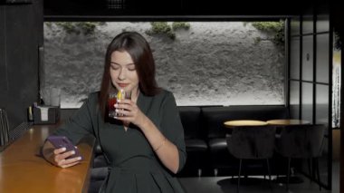 Zarif giyinmiş çekici genç bir kadın şık bir barda canlı bir telefon görüşmesi yaparken canlı bir kokteyl yudumluyor. Modern ortam, şık dekoru ve yumuşak ışıklandırmasıyla, bağlantı atmosferini geliştirir.