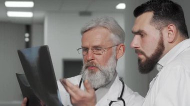 Seçkin sakallı ve gözlüklü kıdemli bir erkek doktor genç meslektaşı ile modern hastane ortamında röntgen taramalarını analiz ediyor. Atmosfer profesyonellik ve işbirliği ile dolu.