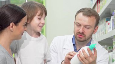 Dost canlısı bir erkek eczacı genç bir anneye ve gülümseyen oğluna parlak ve iyi stoklanmış bir eczanede çeşitli ürünleri keşfederken dikkatle rehberlik eder. Sıcak etkileşim sağlık hizmetlerinde kişiselleştirilmiş hizmetin önemini vurgular,