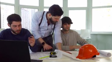 Odaklanmış genç beyaz bir mimar ilgili meslektaşlarına karmaşık proje planlarını tutkuyla açıklıyor. Sahne, modern bir ofis masasında üç inşaat mühendisi yakalıyor, planlar, bir dizüstü bilgisayar ve somutlaşmış bir şapka ile çevrili.
