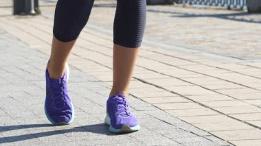 Açık tenli genç bir kadın, dinamik bacağını parıldayan mavi suyla çevrili manzaralı bir nehir yolunda geriyor. Zarif bir şekilde ayaklarını çeviriyor, şık mor spor ayakkabılar giyip tozluklarını giyerken hareketler arasında geçiş yapıyor.