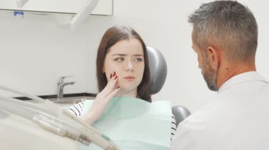 Endişeli bir ifadeye sahip çekici genç bir kadın dişçisine danışırken yanağına hafifçe dokunuyor. Sahne diş bakımının özünü yakalıyor, diş ağrısına değinmenin önemini vurguluyor ve...