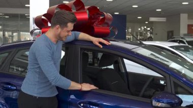 Neşeli genç beyaz bir adam, parlak bir hediye yayı ile süslenmiş parlak mor bir hatchback 'e yaslanırken başparmağını kaldırıyor. Modern bir araba galerisindeki bu canlı sahne heyecan ve onay yayıyor, başarılı bir arabanın heyecanını yakalıyor.