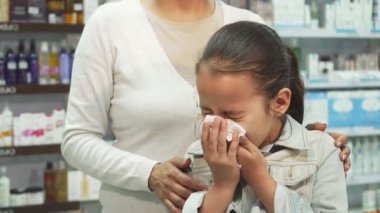 Sıcak bir eczanede, uzun siyah saçlı genç bir kız hapşırır ve burnunu peçeteyle siler, gözle görülür şekilde üzgün görünür. Şefkatli annesi arkasında duruyor, nazikçe omzunu tutuyor, arka planda sevgi ve endişe dolu bir an sergiliyor.