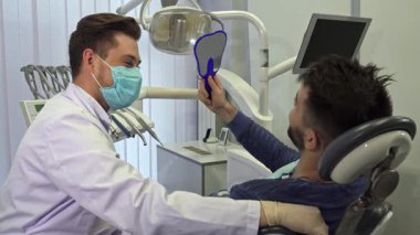 Sakallı yakışıklı esmer bir adam aynaya bakıyor, dişçi muayenehanesinde gülümsemesini değerlendiriyor. Maske takan ve üniforma giyen yetenekli sarışın dişçi yakınlarda oturuyor ve dost canlısı bir tavır takınmaya hazır.