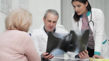 Parlak, modern bir klinikte olgun bir erkek doktor ve genç bayan meslektaşı yaşlı bir hastanın röntgenlerini dikkatle inceliyor. Bu görüşmeler, sağlık sorunlarının teşhis edilmesi ve tedavisinde takım çalışmasının önemini vurguluyor.