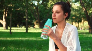 Huzurlu bir parkta, at kuyruklu siyah saçlı güzel bir kadın yoga dersleri arasında ferahlatıcı suyu yudumlarken mutlu bir şekilde gülümsüyor. Bereketli yeşilliklerle çevrili neşeli ifadesi mutluluk ve sükunet yayıyor, sağlıklı bir şekilde vücut buluyor.