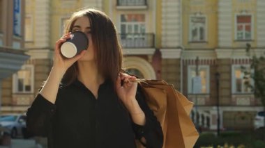 Lezzetli kahverengi saçlı bir kız, canlı bir şehir caddesinde neşe saçarak kahvenin tadını çıkarıyor. Bu şık genç kadın, omzunun üzerinden bir alışveriş çantası sarkıtılmış, zarif bir halk mimarisinin arkaplanına karşı poz veriyor,