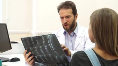 Parlak bir doktor ofisinde, odaklanmış bir doktor genç bir bayana danışırken röntgeni inceler. Atmosfer profesyonelce ama şefkatli, çünkü onun sağlık sorunları ve tavsiyelerini düşünceli bir şekilde tartışıyor.