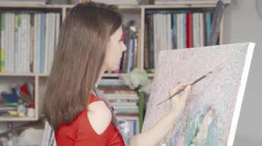 Güzel bir genç kadın, canlı bir sanat atölyesinde canlı bir fırçayla ustalıkla canlı renkler uygulayarak tuvaline derinden odaklanır. İlham verici sanat malzemeleri ve rahat bir stüdyo atmosferiyle çevrelenmiş bir şekilde yaratıcılığı ve tutkuyu somutlaştırıyor.
