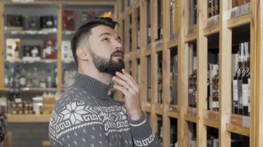 Yakışıklı sakallı bir adam, şık bir kazak giymiş, sıcak, davetkar bir dükkanda enfes şarap şişeleriyle dolu raflara dikkatle bakıyor. Bu sahne kutlamanın ve seçimin özünü yakalıyor. Mükemmel olanı seçmeye hazırlanıyor.