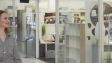 Uçan kızıl saçlı çekici bir kadın şık bir alışveriş merkezinde şık bir mağazaya düşünceli bir şekilde bakıyor. Elinde alışveriş torbalarıyla, tüketim zevkini somutlaştırıyor, moda seçimlerini keşfediyor ve perakende terapisinin canlı atmosferini kucaklıyor.