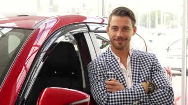 Keyifli bir adam canlı kırmızı arabasının yanında duruyor, büyüleyici bir gülümsemeyle anahtarları gururla sergiliyor. Şık, kareli bir ceket giymiş, neşe ve memnuniyet saçıyor, yeni bir alışverişin heyecanını somutlaştırıyor.