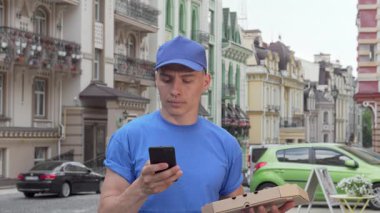 Mavi şapkalı ve gömlekli kendini adamış bir erkek kurye canlı bir şehir caddesinde duruyor. Lezzetli bir pizza teslim etmeye hazırlanırken akıllı telefonunu kontrol ediyor. Elinde bir karton kutuyla, şehir manzarasında geziniyor, koşuşturmayı ve koşuşturmayı somutlaştırıyor.