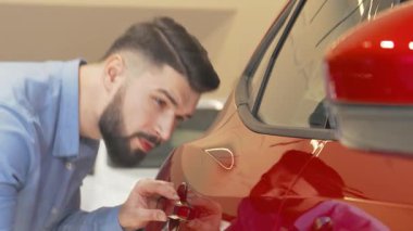 Şık sakallı bir adam modern bir bayilikte parlak kırmızı bir otomobili dikkatle inceler, anlayışlı zevkini gösterir. Bu büyüleyici sahne, lüks bir araç seçmenin heyecanını vurguluyor, stil ve teknolojiyi harmanlayarak canlı bir şekilde