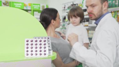Kendini adamış bir eczacı, dikkatli annesi dikkatle izlerken, küçük bir çocuğa enerjik haplar veriyor. Sahne parlak bir eczanede sıcak bir anı yakalıyor, sağlık dolu renkli rafların arasında aile sağlığı ve bakımı vurguluyor.