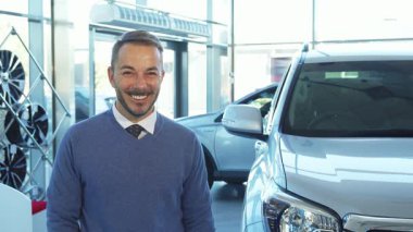 İyi bakımlı sakallı ışıldayan bir adam yeni arabasının anahtarlarını tutuyor ve modern bir araba galerisinde heyecan saçıyor. Şık gri bir kazak ve gevrek beyaz bir gömlek giymişti. Parlak aracın yanında, kendine güvenen bir şekilde duruyordu.