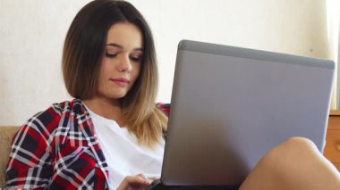 Genç bir kadın rahat bir şekilde rahat bir kanepeye oturur, ara sıra pencereden dışarı bakarken dizüstü bilgisayarına odaklanır. Şık, kareli gömleği ve rahat tavırları sıcak, davetkar bir atmosfer yaratıyor. Üretkenlik ve hayal kurmak için mükemmel.