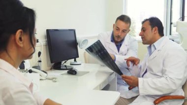 İyi aydınlatılmış bir ofiste, iki erkek doktor röntgen filmlerini dikkatle incelerken, genç bir kadın sabırla sonuçlarını beklerken ciddi bir tartışmaya giriyorlar. Sahne tıbbî profesyonelliğin özünü ve berraklığın önemini yansıtıyor.