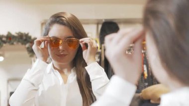 Çekici genç bir kadın şık bir butikte şık güneş gözlüğü denerken neşeyle ışıldıyor. Göz giysisinin canlı renkleri onun neşeli ruhunu yansıtıyor. Alışverişi zevkli bir deneyim haline getiriyor.
