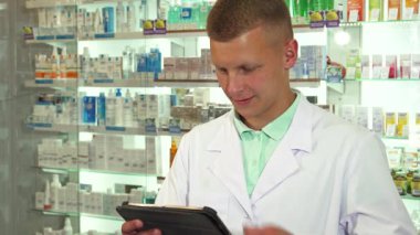 Beyaz tıbbi önlüklü odaklanmış bir erkek eczacı, tablet üzerinde ustalıkla gezinirken telefonla bir hastayla yakınlaşıyor. Düzenli ilaç ürünleri ile çevrelenmiş halde, dolup taşan eczanede profesyonelliği ve kendini adamışlığı simgeliyor.