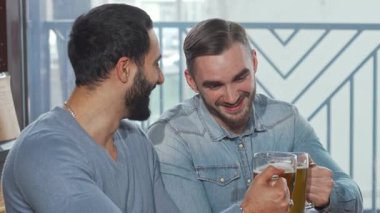 İki neşeli genç adam, biri sakallı ve diğeri şık saç kesimli, buzlu bira bardaklarını canlı bir bar atmosferinde tokuşturuyorlar. Gülüşleri ve yoldaşlıkları, gevşemek için mükemmel bir dostluk ve kutlama sahnesi yaratıyor.