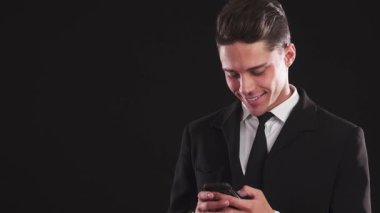 Özel dikim takım elbiseli şık bir genç adam telefonunda kendine güvenen büyüleyici gülüşü var. Siyah arka plana çarpıcı bir profil çizerek, entellektüellik ve cazibe yayıyor, zahmetsizce dikkat çekiyor.