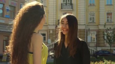 İki şık bayan arkadaş canlı bir şehir caddesinde neşeli bir sohbete girişiyor, bir kız omzunda bir alışveriş torbasını şakayla dinlendiriyor. Sahne pozitiflik saçıyor, arkadaşlığın güzelliğini ve canlı şehir arkaplanını gözler önüne seriyor