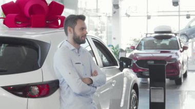 Kendine güvenen bir erkek sürücü, ışıldayan yeni aracının yanında durur, gururla elinde parlak bir gülümsemeyle arabanın anahtarlarını tutar. Parlak sergi salonu, arabanın üzerinde büyük kırmızı bir fiyonk ile süslenmiş, heyecan ve yeni sahiplik heyecanı yayıyor.