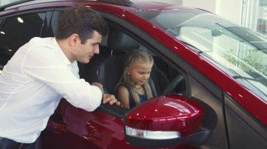 Şefkatli bir baba açık pencereden sarkmış, kızına arabaların düğmelerini açıklarken, kızına direksiyon başında güven içinde oturuyordu. Gösterişli araba galerisindeki neşeli etkileşimleri sıcaklık ve bağlantı yayıyor.