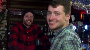 Genç beyaz bir adam, soğuk bir bardak bira kaldırırken arkadaşlarıyla kalabalık bir barda kutlama yaparken neşeyle ışıldıyor. Arka planda, sakallı arkadaşı kahkahaları paylaşıyor, yoldaşlık ve eğlenceyle dolu canlı bir atmosfer yaratıyor.