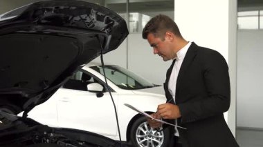Koyu renk saçlı, bakımlı sakallı, odaklanmış bir araba satıcısı, belgelerine bakarken motoru kontrol ediyor. Gösterişli, modern sergi salonunda arka planda el değmemiş beyaz bir araba var.