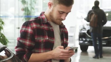 Şık sakallı genç bir adam akıllı telefonuna odaklanmış, parlak araçlarla dolu modern bir araba galerisini keşfederken mesajlar yazıyor. Bu canlı sahne, teknoloji ve otomotiv hayallerinin gerçek olduğu çağdaş tüketiciliğin özünü yansıtıyor.