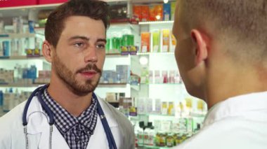 Gıcır gıcır bir tıbbi elbise ve steteskop giymiş odaklanmış bir erkek doktor hastasını dikkatle dinliyor ve şefkatli bir tavır sergiliyor. Çeşitli ilaçlarla dolu renkli raflarla çevrelenen sahne, sağlık hizmetlerinin ve