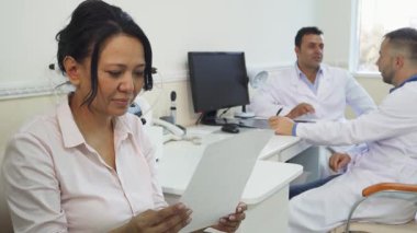 Parlak bir doktor ofisinde, genç bir kadın sağlık raporunu incelerken mutlulukla ışıldıyor ve randevudan sonra memnuniyetini sergiliyor. Sahne onun neşesini yakalıyor, dikkatli erkek doktorların tartışması altında.