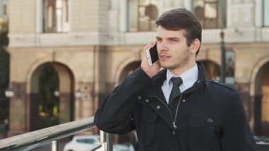 Keskin takım elbiseli genç bir adam dışarıda duruyor, ciddi bir telefon görüşmesi yapıyor. İfadesi, çalkantılı bir şehir zemininde önemli bir iş görüşmesini yönetirken azim ve endişenin bir karışımını ortaya koyuyor.