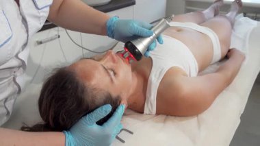 Yetenekli bir güzellik uzmanı, dingin bir güzellik salonundaki tedavi masasında rahatlayan olgun bir kadına gençleştirici bir RF kaldırma tedavisi uyguluyor. Gelişmiş radyogereksinim cihazı derisinin üzerinde süzülerek kolajen üretimini teşvik ediyor.