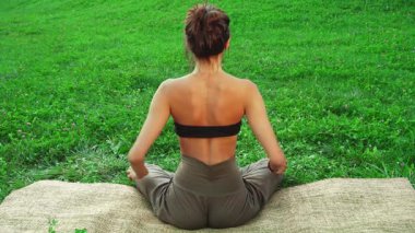 Genç bir kız doğal yoga minderinin üzerinde huzur içinde oturur. Sırtı izleyiciye dönük bir şekilde yoga pozu verir. Etrafı yemyeşil çimlerle çevrili, sakin ifadesi ve rahat duruşu doğadaki sükunet ve farkındalığın özünü temsil ediyor.