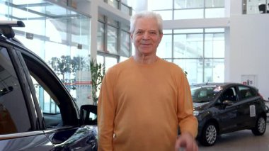 Neşeli, kıdemli, beyaz bir adam parlak bir galeride elinde yeni araba anahtarlarıyla gururla duruyor. Şık araçlarla dolu canlı sergi salonu, yeni alımını kutlarken heyecanını ve memnuniyetini yansıtıyor.