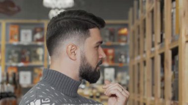 Yakışıklı sakallı bir adam şık bir süpermarkette özenle şarap seçerken görülüyor. Kendine güveni ve günlük süveteri rahatlamış bir alışveriş deneyimini yansıtıyor. Mükemmel şişeyi seçme zevkini vurguluyor.