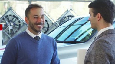 Sakallı ve koyu renk saçlı keyifli bir adam çarpıcı yeni arabasının anahtarlarını iyi giyimli bir satıcıdan alıyor. Yeni maceralara atılmaya hazır anahtarlarını gururla sergilediği için havadaki heyecan hissediliyor.