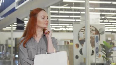 Neşeli bir kadın canlı alışveriş merkezini keşfederken elinde alışveriş torbalarıyla kameraya ışıldıyor. Şık kıyafeti ve ışıl ışıl gülüşü büyük anlaşmaları ve moda modasını keşfetmenin heyecanını yansıtıyor.