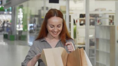 Uçan kızıl saçlı mutlu bir kadın alışveriş merkezinin canlı ortamında neşe saçan alışveriş çantasına bakıyor. Neşeli gülüşü ve şık kıyafeti renkli vitrinlerle çevrili başarılı bir alışveriş çılgınlığının heyecanını yansıtıyor.
