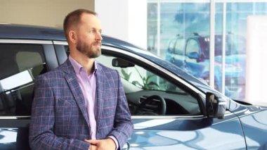 İyi bakımlı sakallı karizmatik bir adam modern bir otomobil galerisinde gösterişli bir arabanın yanında gururla durur. Anahtarları tutarken ve başparmak kaldırırken parlak bir gülümseme gösteriyor, araçlara olağanüstü bir güven ve coşku yayıyor.