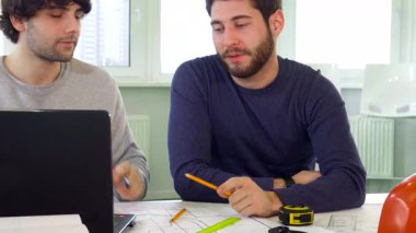 İki yetenekli mimar, biri şık sakallı ve diğeri dağınık saçlı, bir dizüstü bilgisayar ekranını incelerken canlı bir tartışmaya giriyorlar. Sahne, odaklanmış ifadelerini ve mimari aletlerle dolu canlı çalışma alanını yakalıyor.