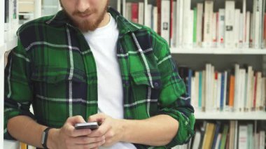 Koyu saçlı ve sakallı genç bir adam renkli kitaplarla dolu canlı bir kütüphanede cep telefonuyla neşe içinde yazı yazıyor. Rahatlamış tavrı ve gülüşü pozitiflik saçıyor, çalışma ortamını davetkar ve canlı hissettiriyor.